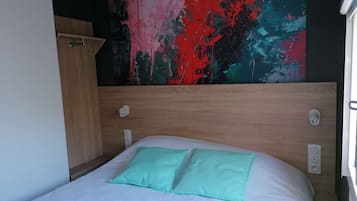 Un lit simple avec un matelas blanc et deux oreillers verts, une tête de lit en bois et un mur orné d’un tableau abstrait aux couleurs vives.