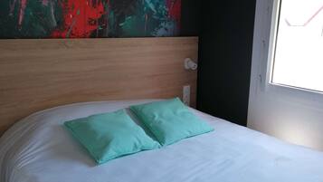A bed with white sheets and two teal pillows, a wooden headboard, and a colorful abstract painting on the wall.