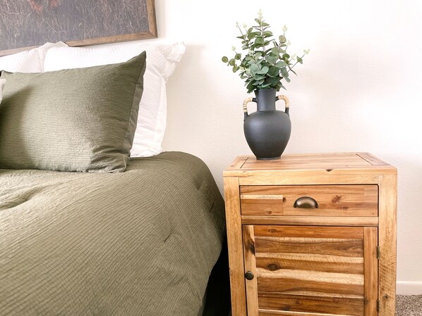 Una cama con una colcha y almohadas verdes, una mesita de noche de madera con un jarrón de plantas verdes, y una pared blanca con un cuadro enmarcado.
