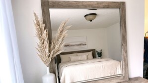Ein Schlafzimmer mit einem großen Spiegel, einem Bett mit weißer Bettwäsche, einer Vase mit Trockenpflanzen und einem Nachttisch aus Holz.