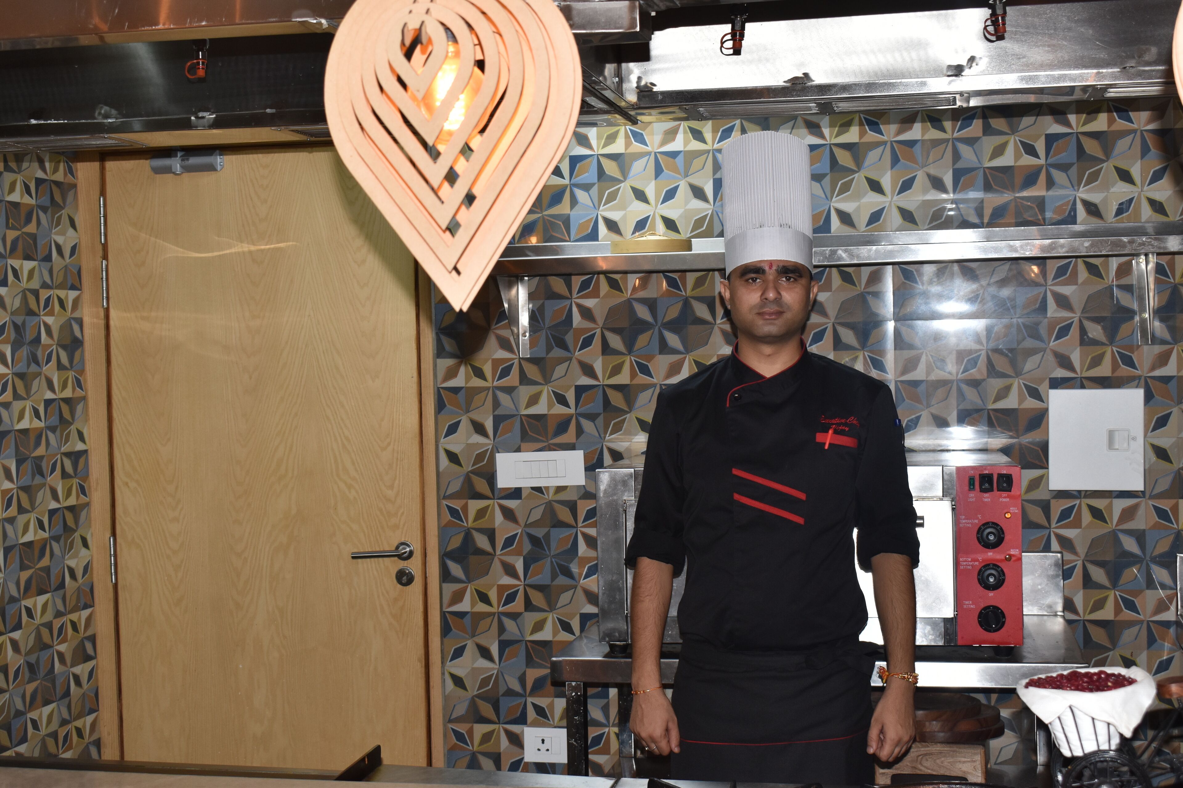 A chef in a professional kitchen with patterned tile walls, stainless steel appliances, and a wooden door.