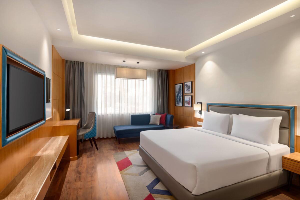 A modern hotel room with a large bed, a seating area with a sofa and armchair, a flat-screen TV, and a large window with curtains.