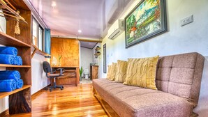 Ein Zimmer mit Holzboden, einer Couch mit Kissen, einem Schreibtisch mit Stuhl und einer Wandklimaanlage.