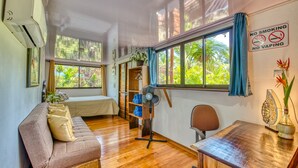 Ein Zimmer mit Holzboden, einer Couch, einem Tisch und einem Stuhl. Es gibt ein Fenster mit Vorhängen, einen Ventilator und ein Schild, das Rauchen und Dampfen verbietet.