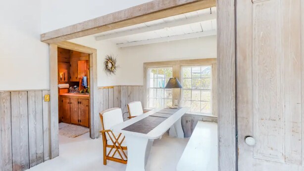 Een eetgedeelte met een lange tafel, witte stoelen en een lamp. Een keuken is zichtbaar door een open deur.