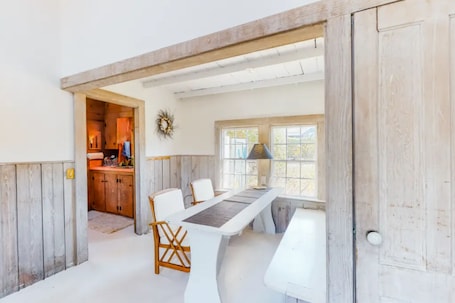 A dining area with a long table, white chairs, and a lamp. There is a kitchen visible through an open door.