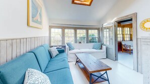 Een gezellige woonkamer met een blauwe bank, een houten salontafel en een witte zithoek. Boven de bank is een dakraam en er is een raam met uitzicht op groen buiten.