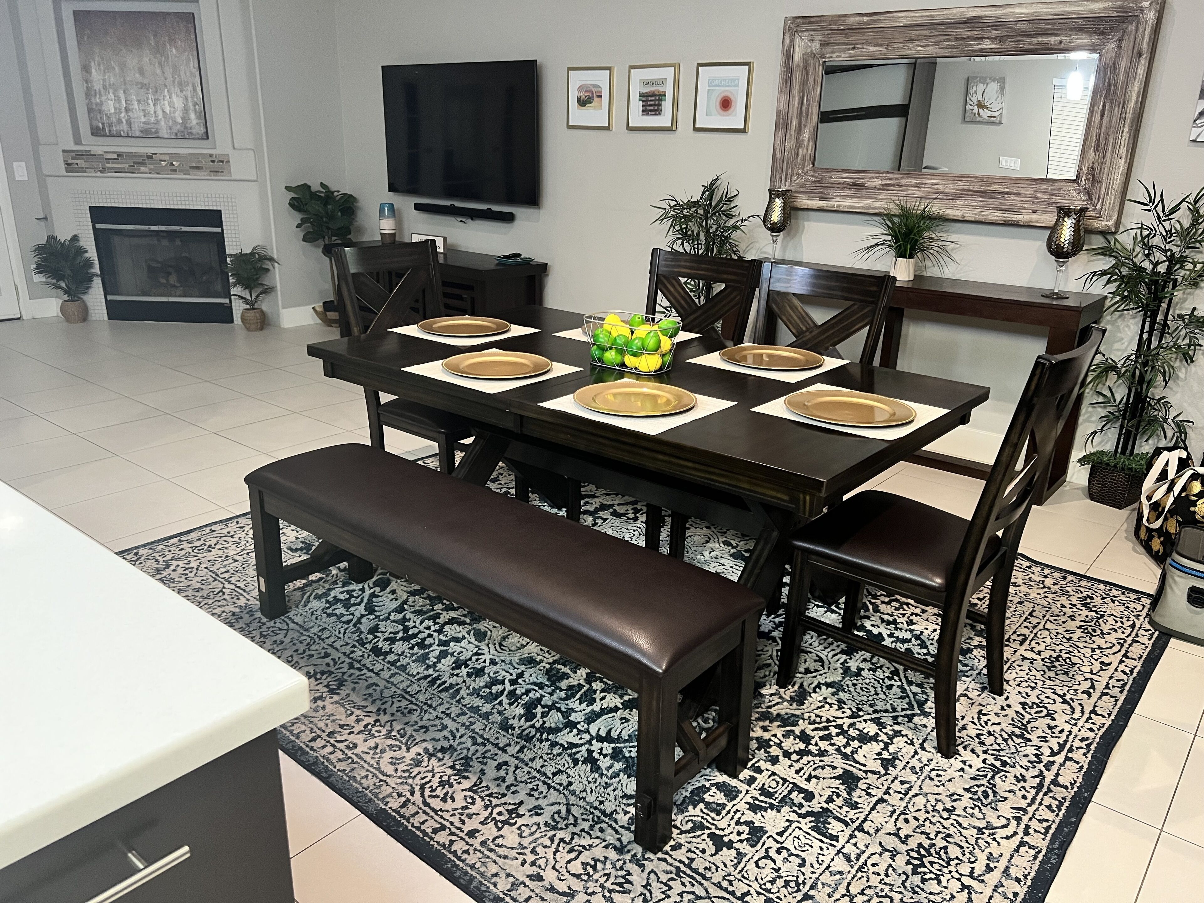 A dining area with a dark wooden table set for six, surrounded by chairs. A bench is placed in front of the table. The room has a tiled floor, a patterned rug, and is decorated with potted plants.