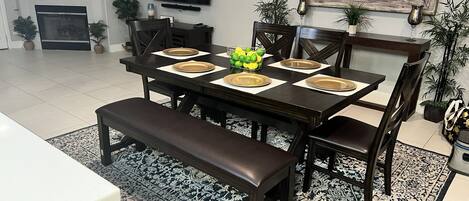 A dining area with a dark wooden table set for six, surrounded by chairs. A bench is placed in front of the table. The room has a tiled floor, a patterned rug, and is decorated with potted plants.