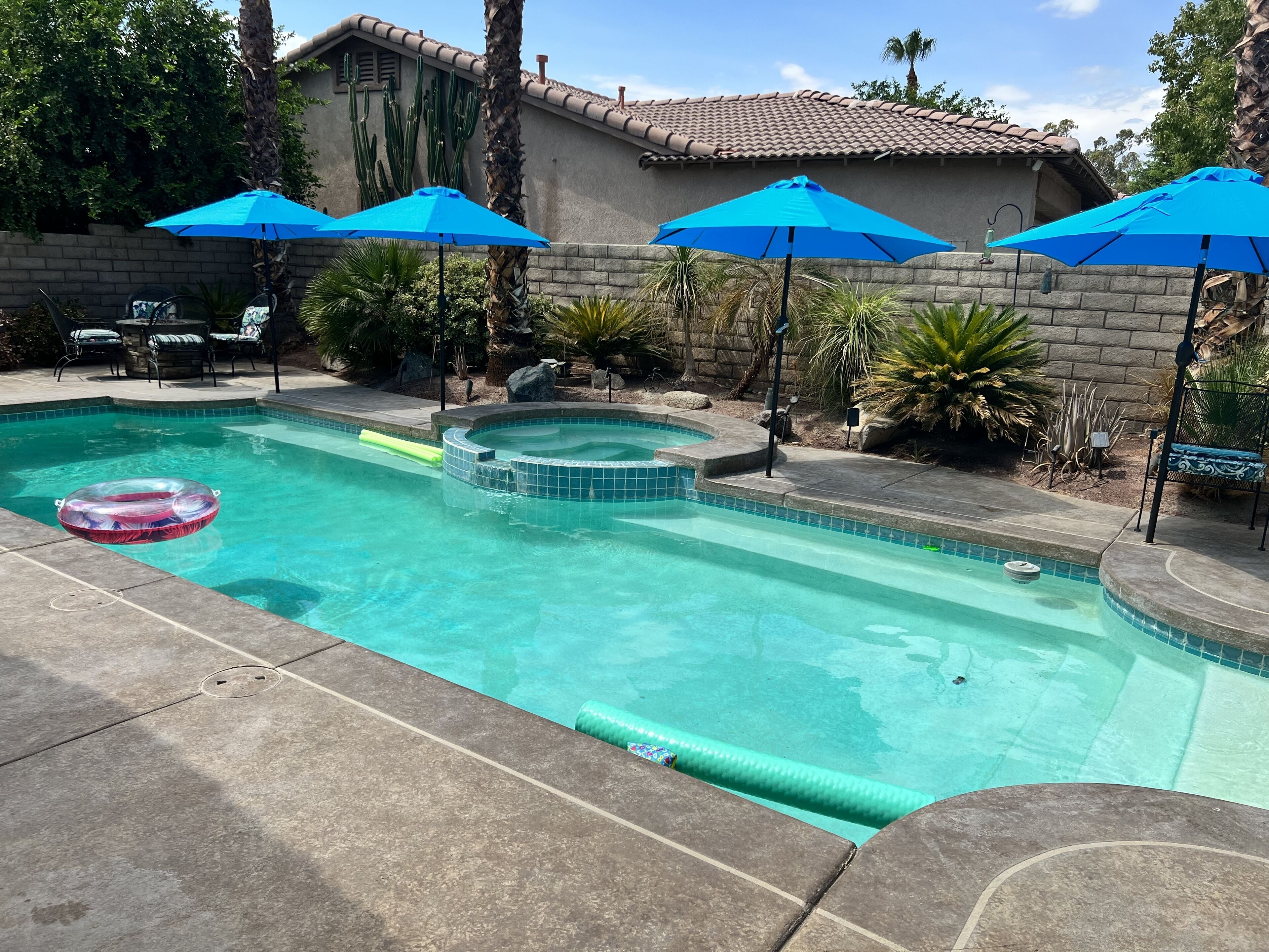 A swimming pool with a hot tub, surrounded by palm trees and blue umbrellas.