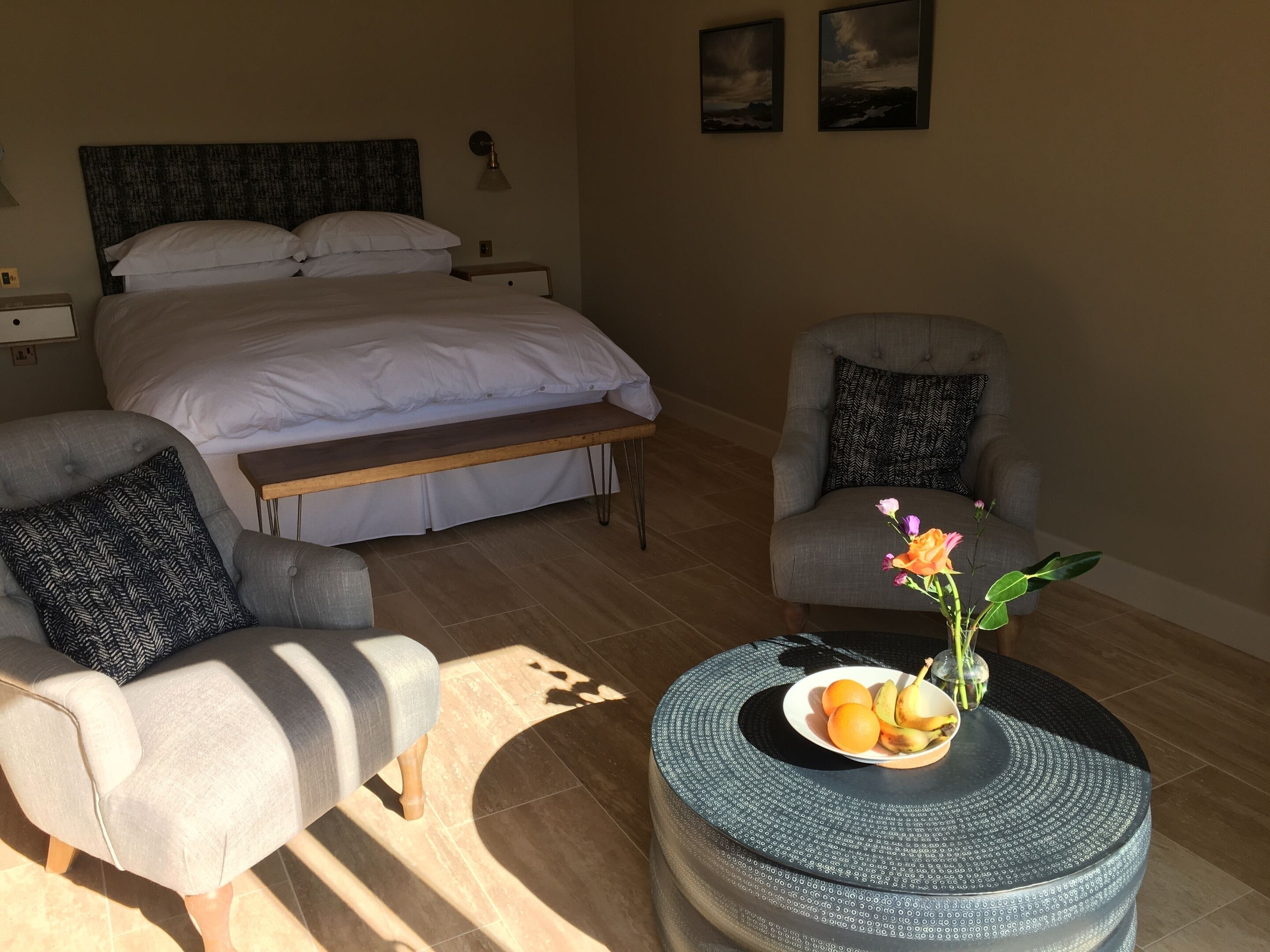 A bedroom with a bed, two armchairs, a bedside table, a round coffee table with fruit, and a wall-mounted lamp.