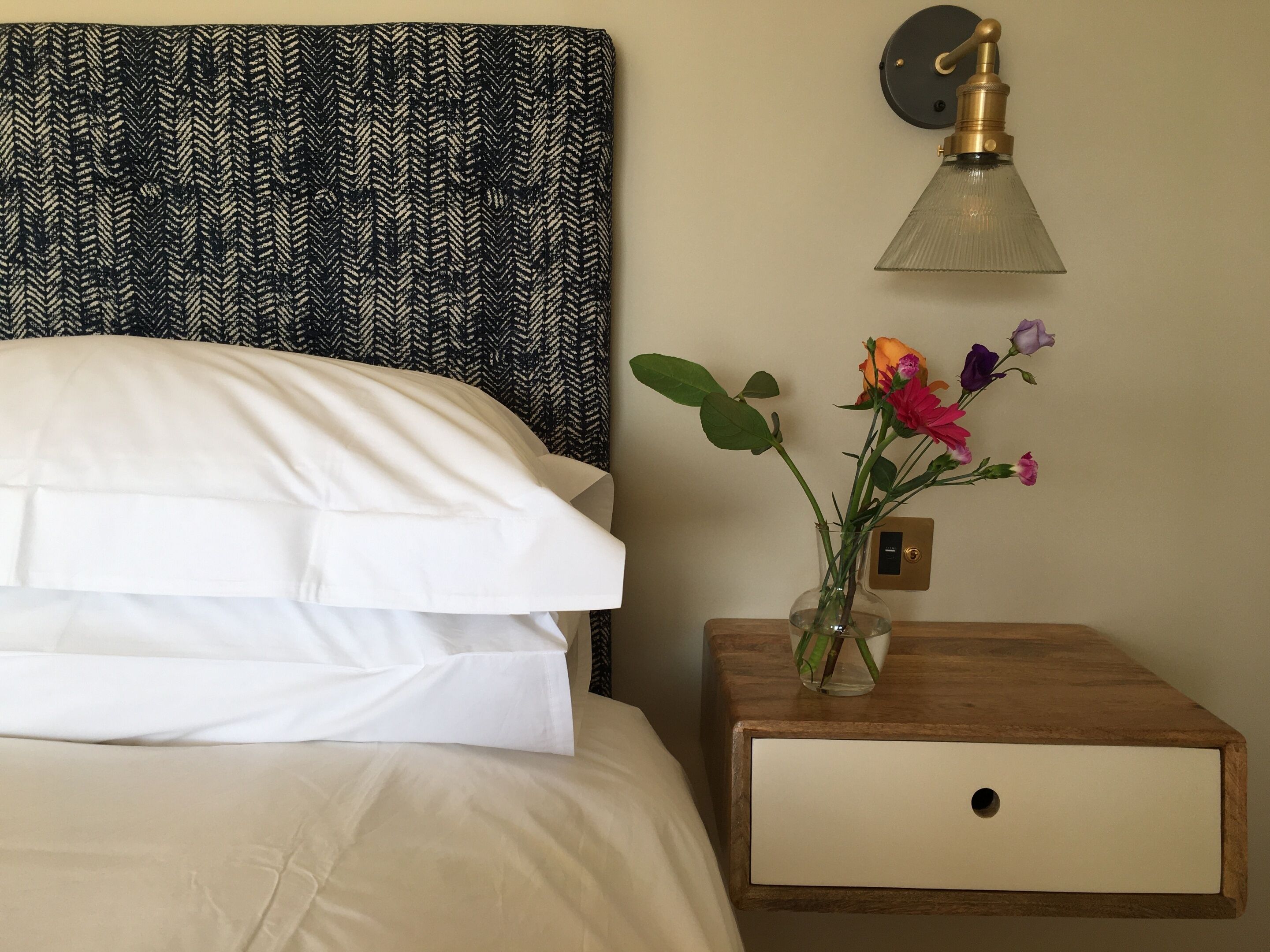 A neatly made bed with white linens, a wooden nightstand with a vase of flowers, and a wall-mounted lamp.