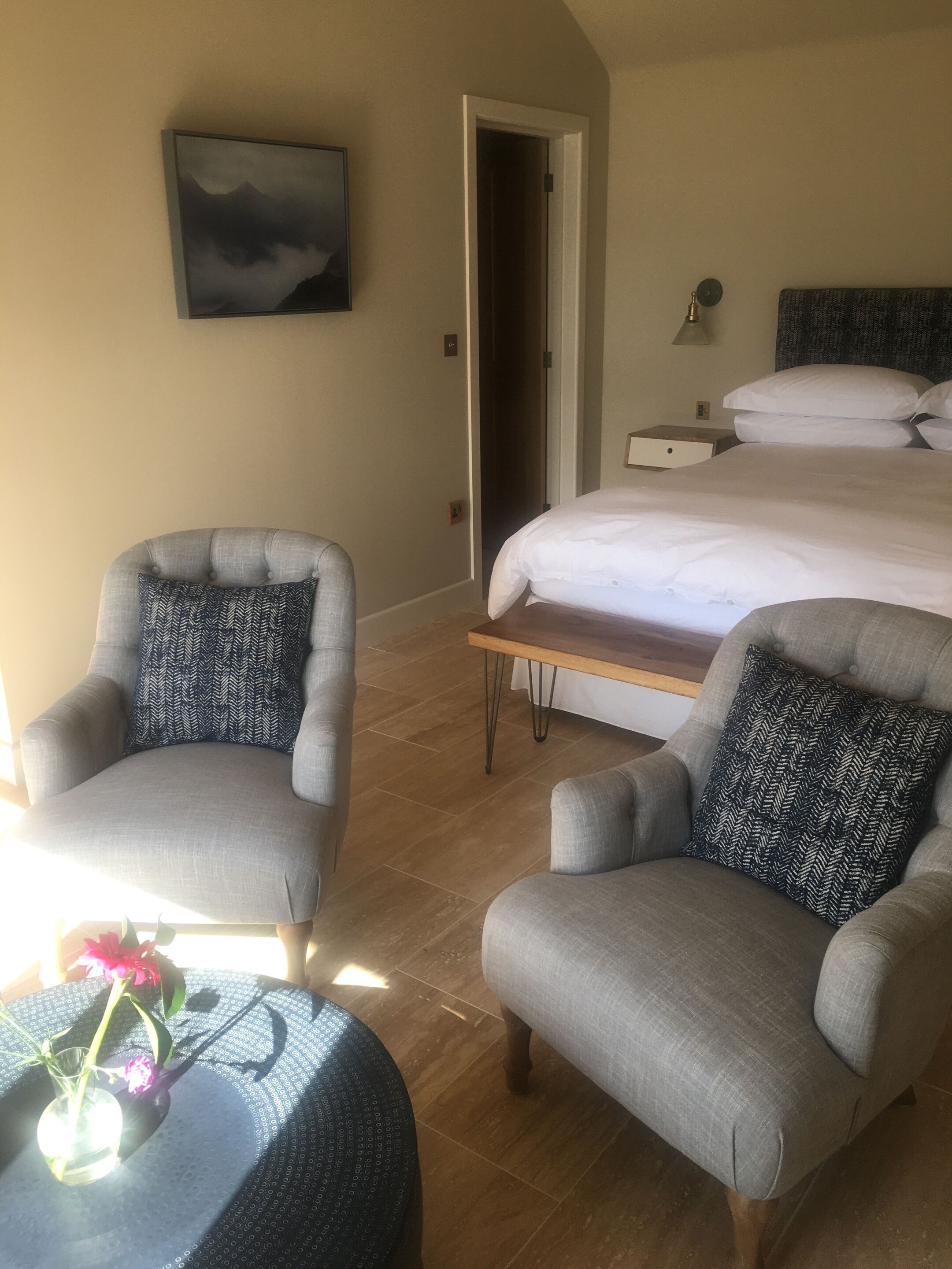 A hotel room with a bed, two armchairs, a small table with a vase of flowers, a painting on the wall, and a door leading to another room.