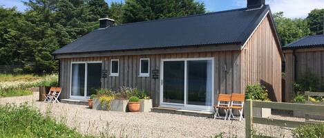 A wooden cabin with a dark roof, large sliding glass doors, and a gravel driveway.
