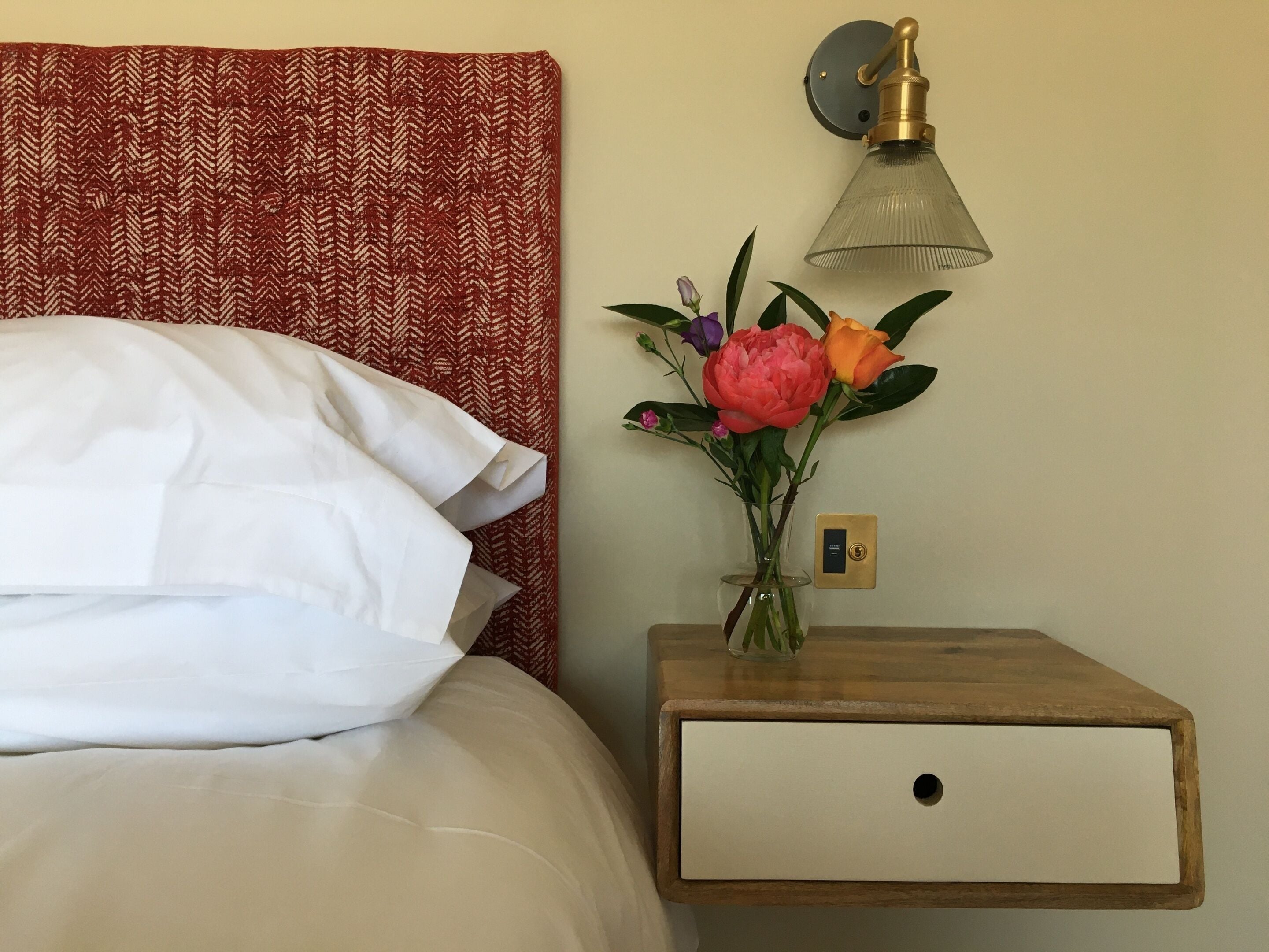 Una cama bien hecha con ropa blanca, una mesita de noche de madera con un florero y una lámpara de pared.
