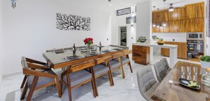 Uma sala de jantar moderna com mesa de madeira e cadeiras estofadas em cinza, cozinha com armários de madeira e um ventilador de teto.