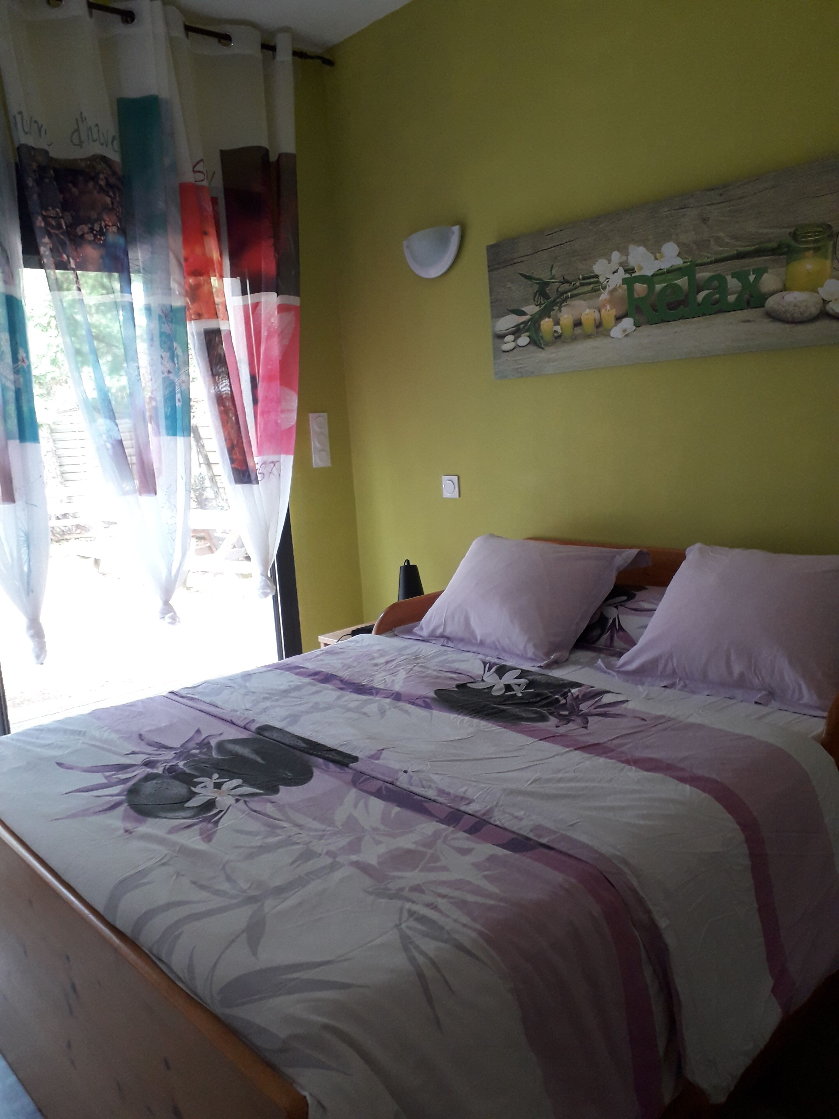 A bedroom with a bed, a nightstand, a wall-mounted lamp, and a window with curtains.