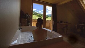 A room with a large bathtub, a wooden ceiling, and a view of a green landscape through a glass door. - AGRITURISMO SAN GALLO (Bormio)