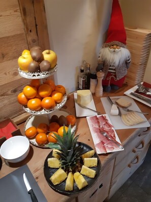 A wooden table with a variety of fruits, including oranges, apples, and pineapples, and some food items like cheese and ham. - AGRITURISMO SAN GALLO (Bormio)