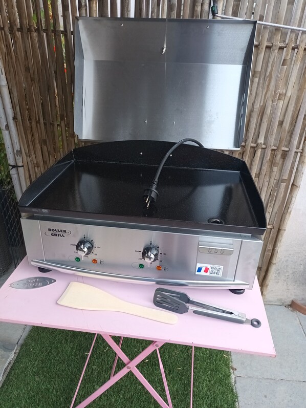 Een rollengrill met bedieningsknoppen en een digitaal scherm, geplaatst op een roze tafel met een spatel en tangen.