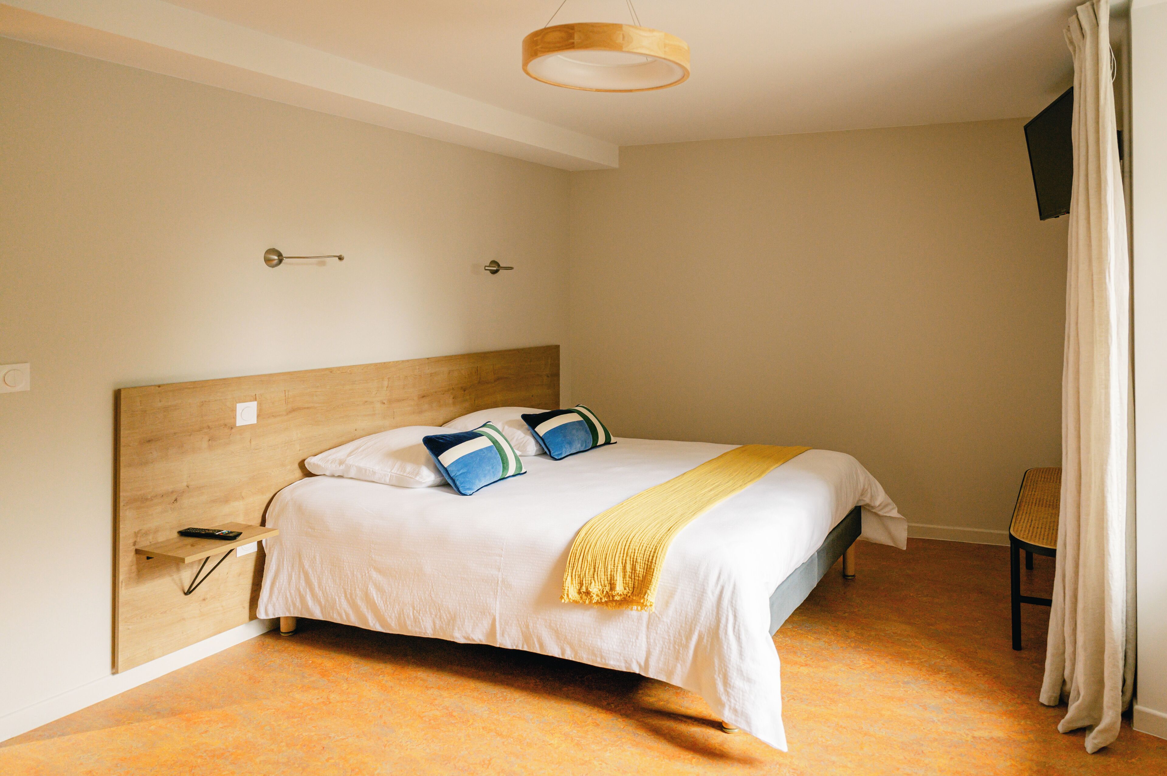 Ein Hotelzimmer mit hölzernem Kopfteil, einem Bett mit weißer Bettwäsche und blauen Kissen, einer gelben Tagesdecke, einem kleinen Nachttisch mit Fernbedienung und einer schlichten Wand mit einer Leuchte.
