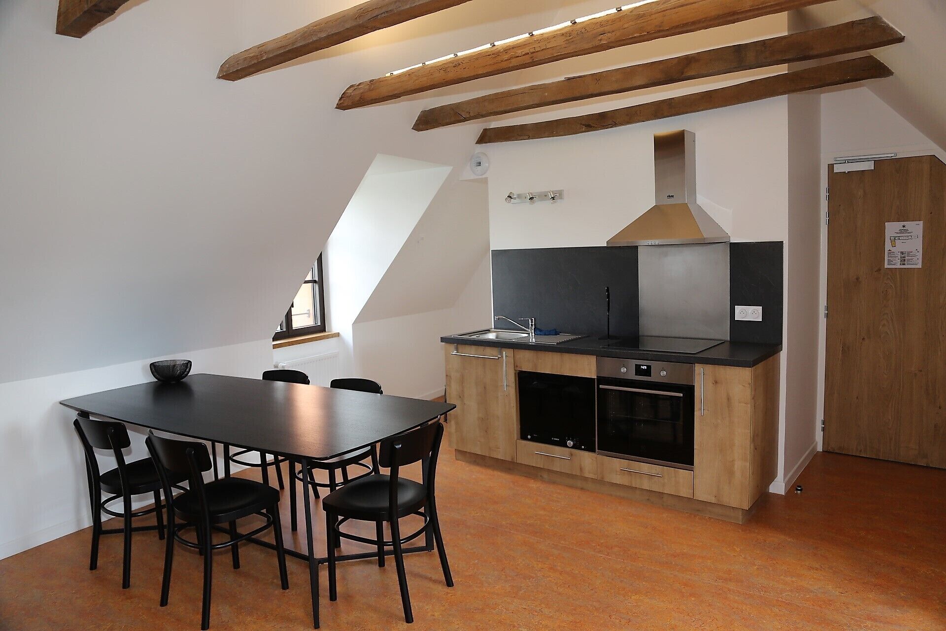 A modern kitchen with a black countertop, wooden cabinets, and a built-in oven. There is a dining table with four chairs and a window providing natural light.