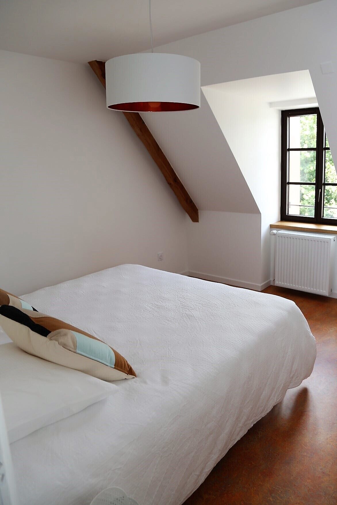 Et soveværelse med skrå væg, seng med hvid madras og pude, et vindue med udsigt til grøn vegetation, og en radiator.
