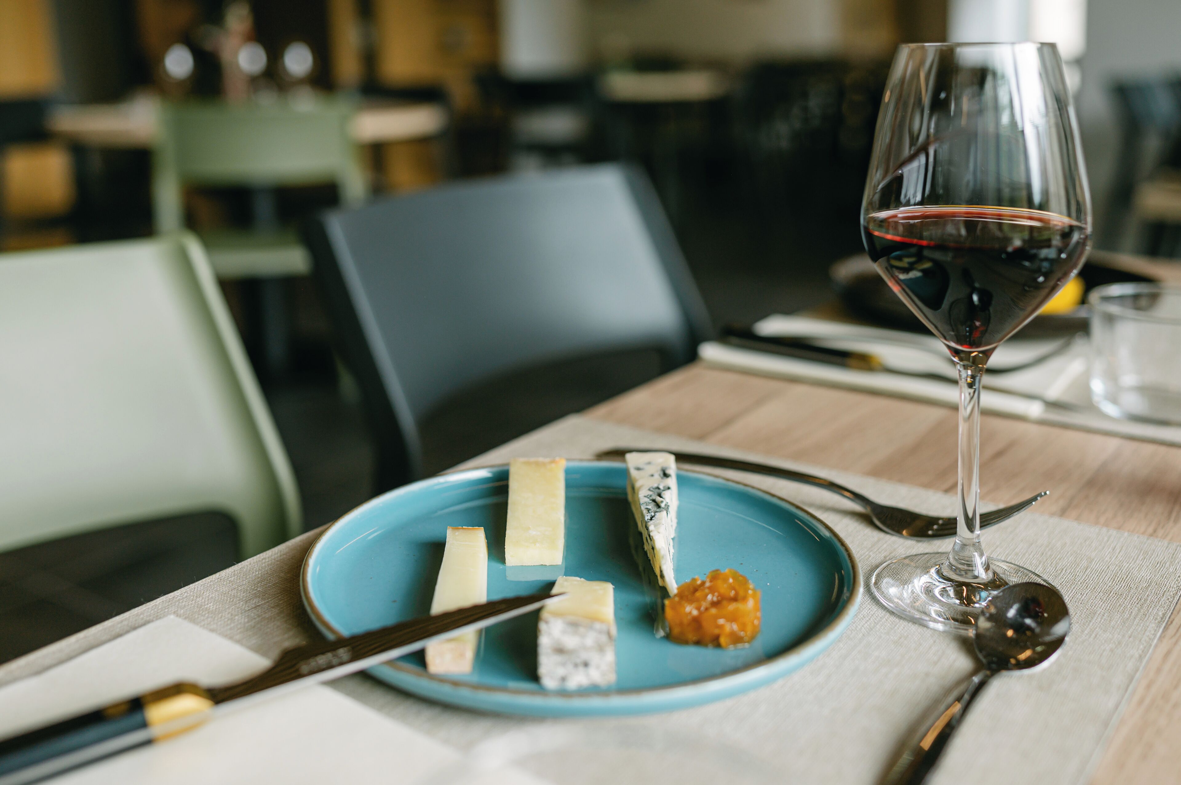 A table with a plate of cheeses, a glass of red wine, and cutlery.