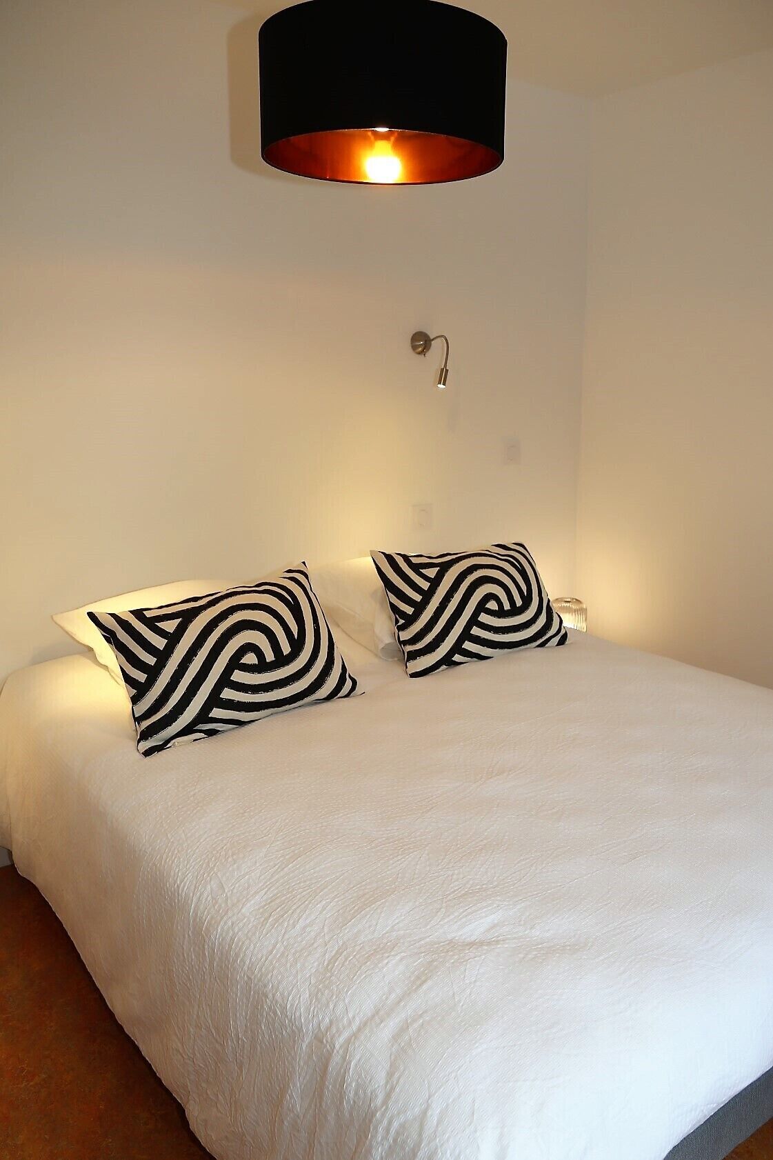 Una cama bien tendida con dos almohadas estampadas, una lámpara negra encima y una lámpara de pared.