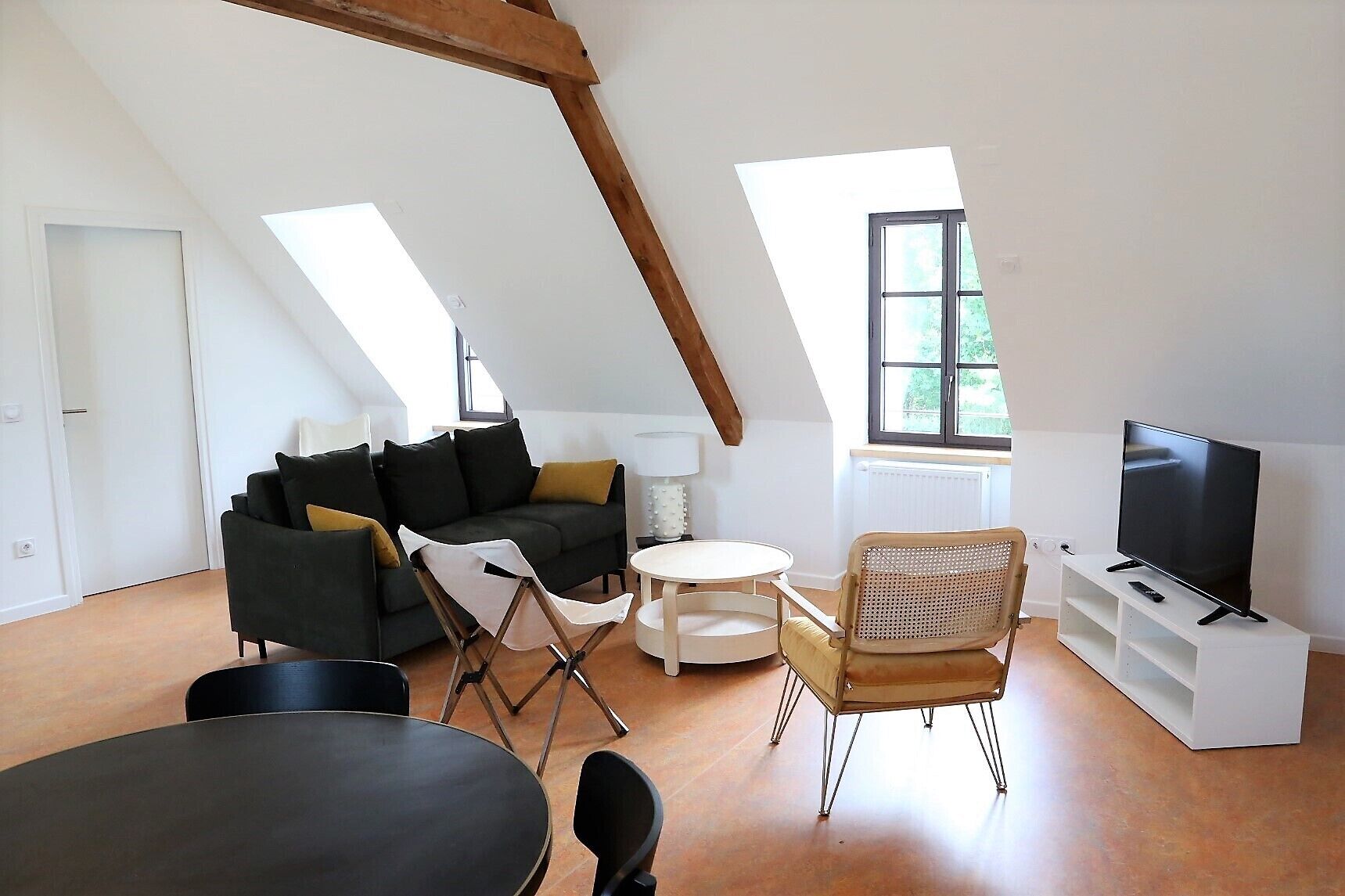 Un salon de style industriel, avec un plafond haut, un canapé noir, une petite table ronde, une chaise pliante, un téléviseur posé sur un meuble blanc et une fenêtre donnant sur un paysage verdoyant.