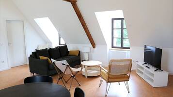 Un salon de style industriel, avec un plafond haut, un canapé noir, une petite table ronde, une chaise pliante, un téléviseur posé sur un meuble blanc et une fenêtre donnant sur un paysage verdoyant.