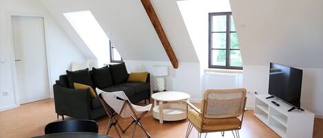 A loft-style living room with a high ceiling, a black sofa, a small round table, a folding chair, a television on a white stand, and a window with a view of greenery.