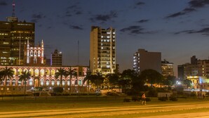 Un paisaje urbano al anochecer con edificios iluminados, incluyendo una estructura histórica destacada con arcos y un rascacielos moderno.
