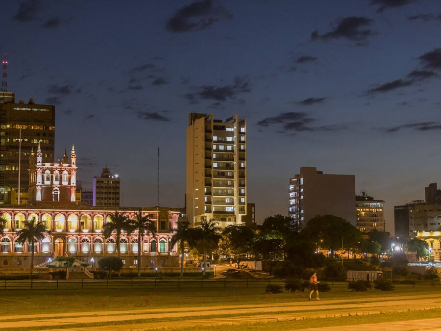 Un paisaje urbano al anochecer con edificios iluminados, incluyendo una estructura histórica destacada con arcos y un rascacielos moderno.