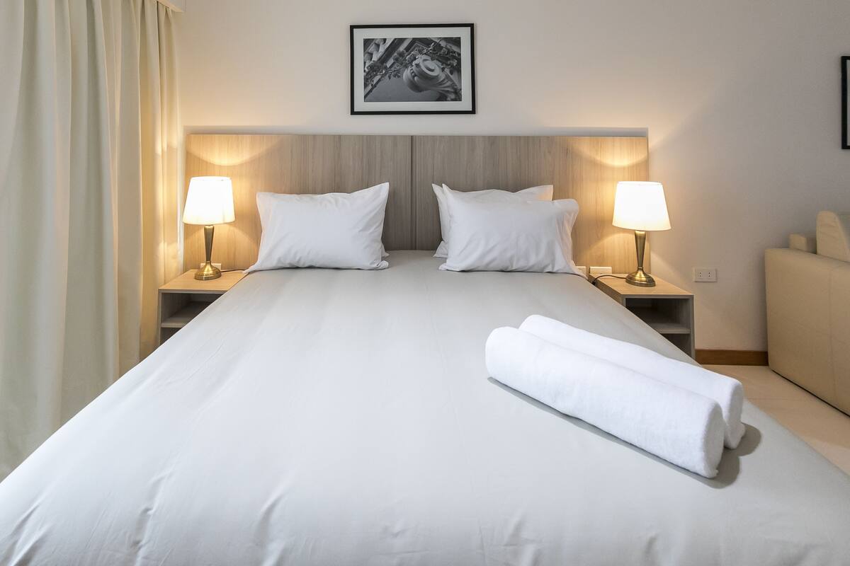 Quarto de hotel com uma cama grande, duas luminárias de cabeceira, uma cabeceira e uma toalha enrolada na cama.