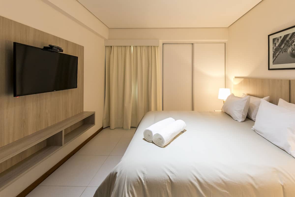 Quarto de hotel com televisão de tela plana na parede, cama com roupa branca, abajur e quadro emoldurado na parede.