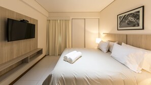 A hotel room with a flat-screen TV mounted on the wall, a bed with white linens, a bedside lamp, and a framed picture on the wall.