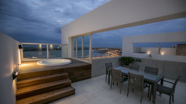 Terraza en la azotea con jacuzzi, mesa de comedor, sillas y vista a la ciudad al anochecer.