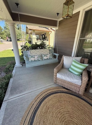 A porch with a wicker chair and a bench, a hanging lantern, and a view of a house in the background.