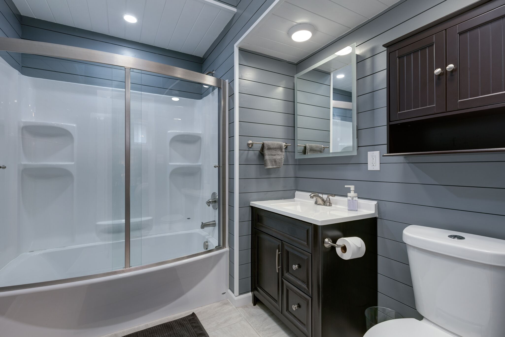 A modern bathroom with a glass-enclosed shower, a white sink, dark cabinets, and a large mirror.