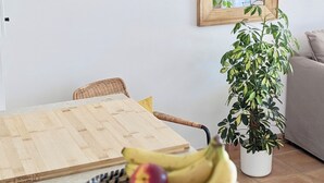 Un coin repas avec une table en bois, des chaises en osier, un panier de fruits contenant des bananes et des pommes, une plante en pot et un miroir fixé au mur.