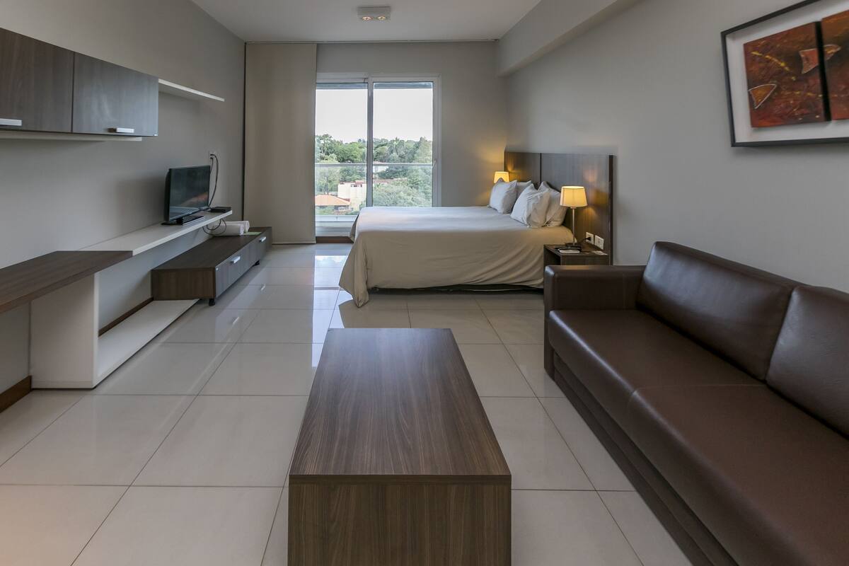 A modern hotel room with a bed, a sofa, a TV, a desk, and a large window.