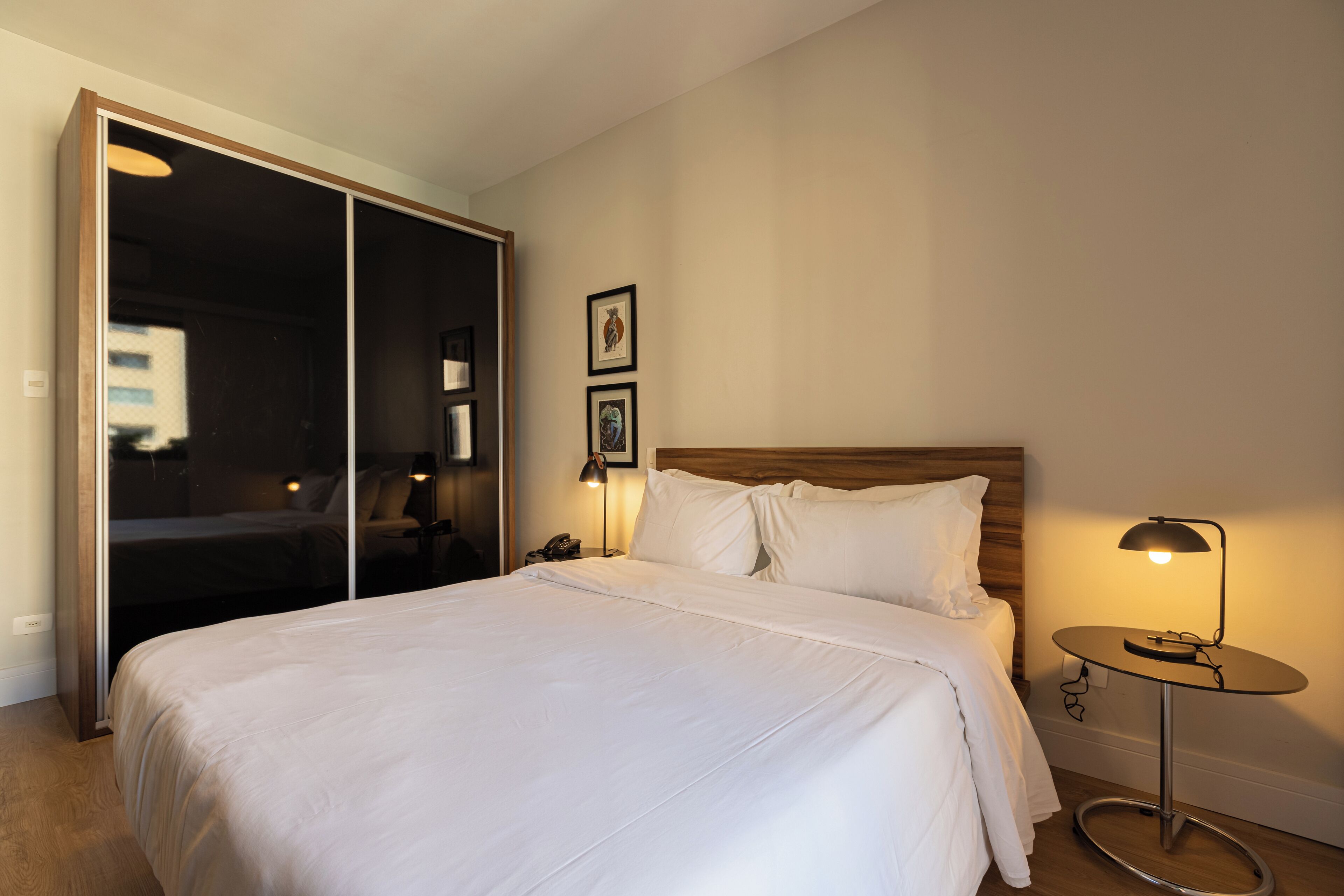 Un dormitorio con una cama grande, una mesita de noche con lámpara, un armario y dos cuadros en la pared.