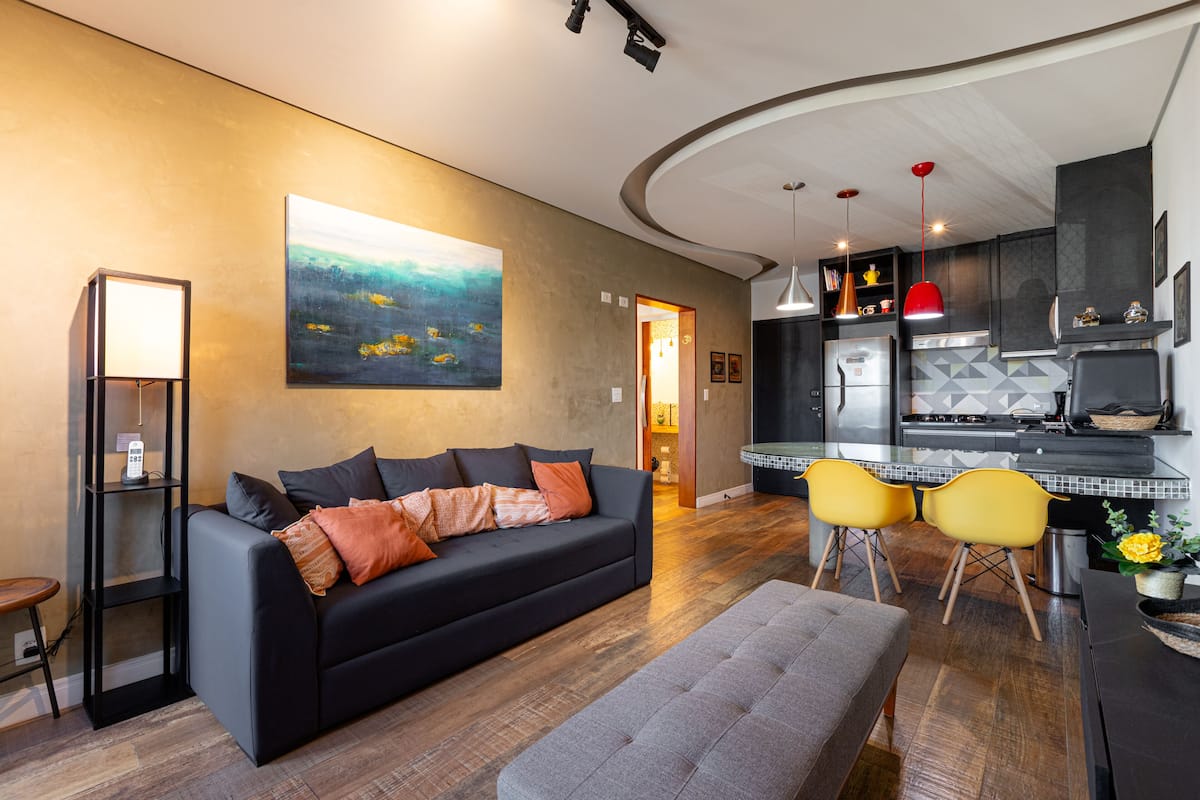 Een moderne woonkamer met een zwarte bank, een grijze poef en gele stoelen. Aan de muur hangt een schilderij, en er is een keuken met een rode hanglamp.