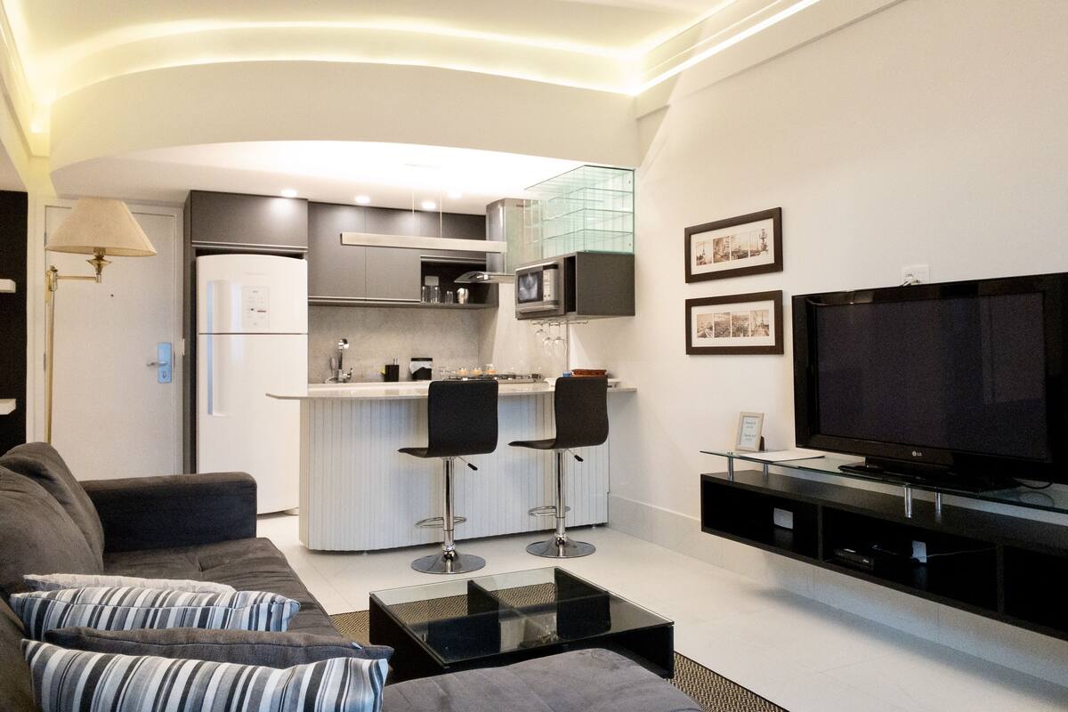 Een moderne woonkamer met een bank, een salontafel, een televisie en een keukenhoek met een koelkast, magnetron en barstoelen.