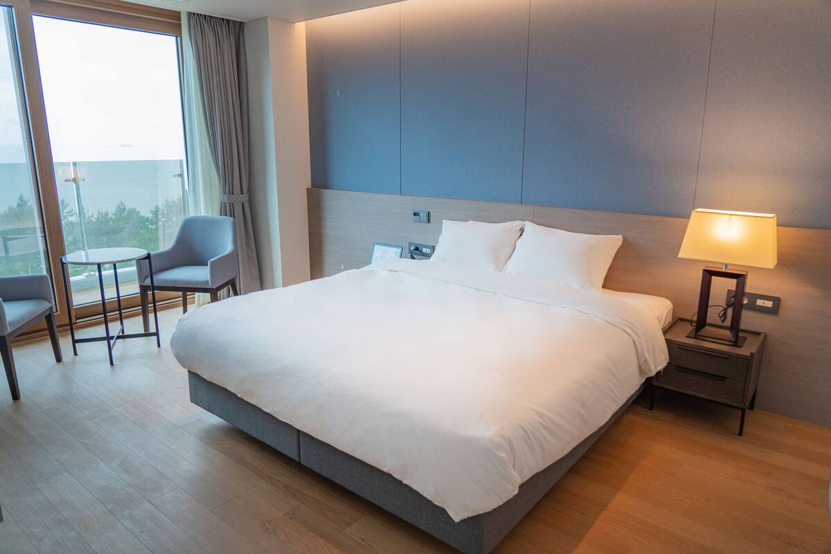 Una habitación de hotel moderna con una cama grande, una mesita de noche con lámpara, una silla y vistas al exterior a través de un ventanal amplio.