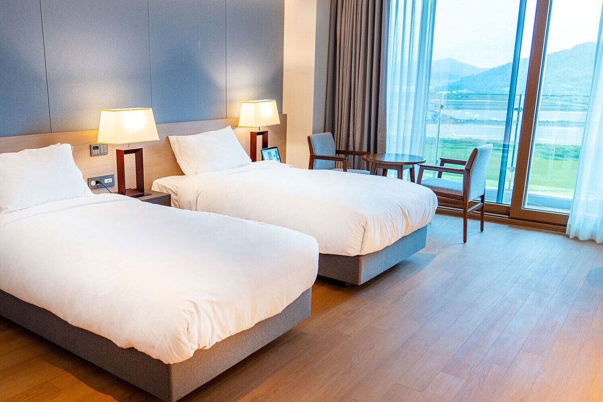 Ein Hotelzimmer mit zwei Betten, Nachttischlampen, einem Sessel, einem kleinen Tisch und Blick auf eine Landschaft durch große Fenster.