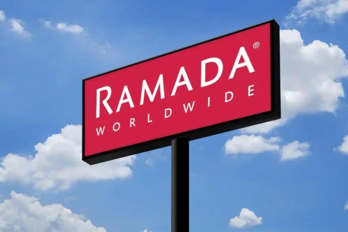 Un letrero rojo con la inscripción 'RAMADA WORLDWIDE' sobre un cielo azul con nubes.