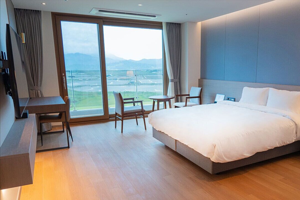 Ein Hotelzimmer mit einem großen Bett, einem Schreibtisch, Stühlen, einem kleinen Tisch, einer Lampe und Blick ins Freie.