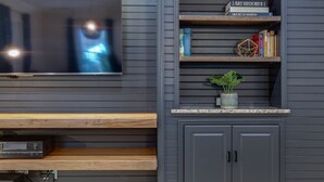 Een moderne woonkamer met een flatscreen televisie aan de muur, houten planken met boeken en decoratieve objecten, en een kleine potplant op een aanrecht.